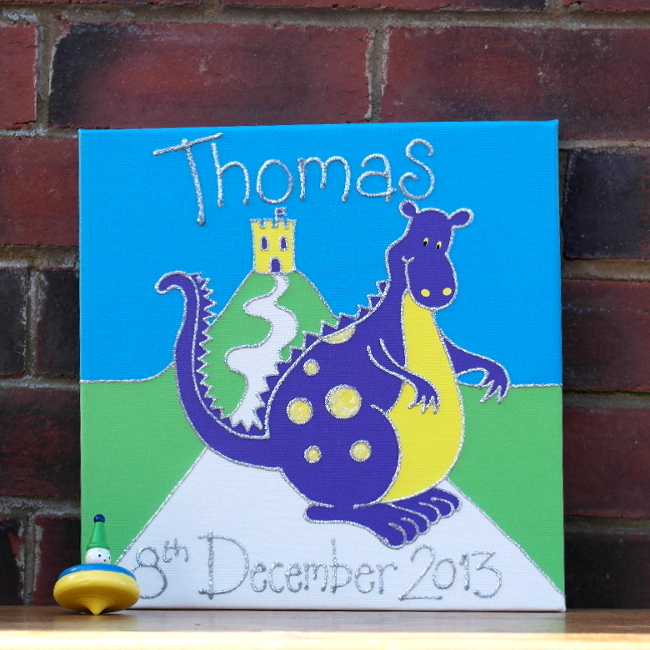 A personalised canvas picture complete with childs name and date of birth in silver glitter. The canvas picture shows a huge purple dragon with yellow spots and a yellow tummy standing in front of a castle on a hill.