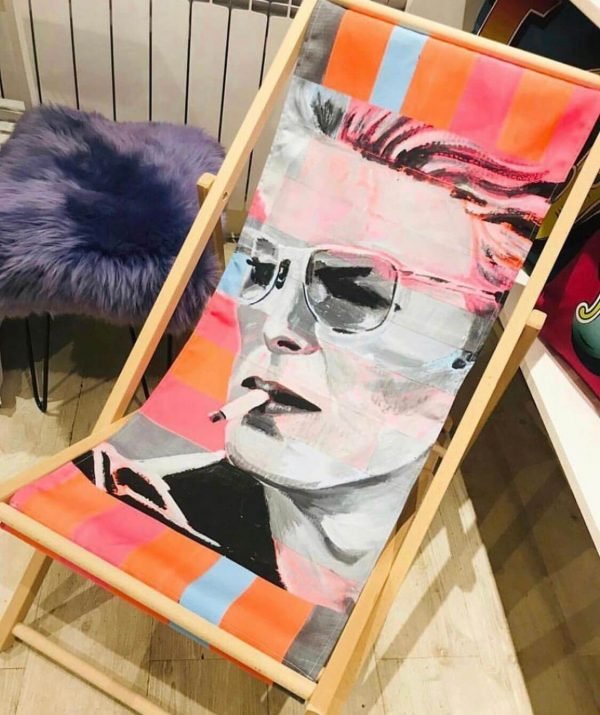 A neon bowie deck chair with a wooden frame, and a canvas sling seat with a David Bowie design printed on it.