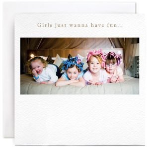 A white square card with a photographic image of 4 little girls having a pamper day with curlers in their hair, lying on a bed.