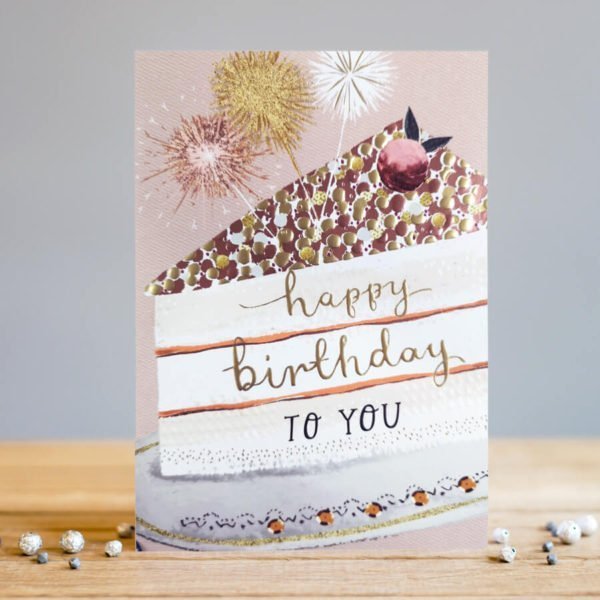 A rectangular card with a dusky pink background with an image of a slice of birthday cake which has sparklers on the top of it. The words Happy Birthday to you are printed on the layers of the cake.