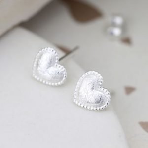 A pair of silver heart studs earrings, which have tiny beading around the edges and the inset section is a brushed or scratched finish.