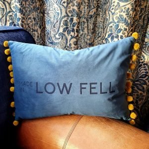 An indigo blue luxurious velvet cushion with olive green pom pom trim. The cushion has been engraved with MADE IN LOW FELL