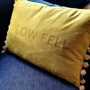 An olive green luxurious velvet cushion with buttermilk pom pom trim. The cushion has been engraved with MADE IN LOW FELL