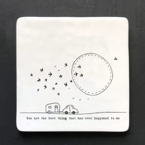 A white square coaster with an image of a caravan with birds and the sun in the sky