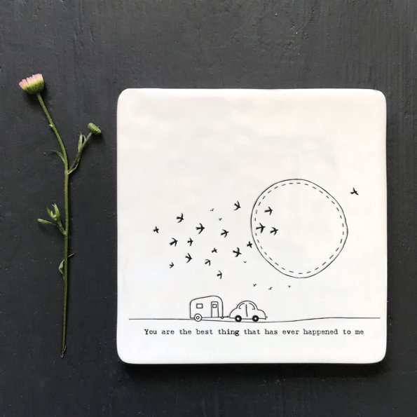 A white square coaster with an image of a caravan with birds and the sun in the sky