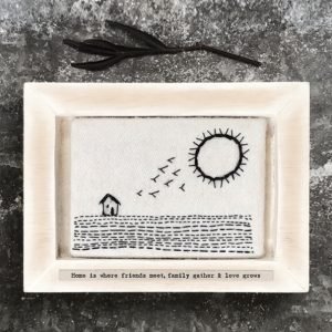 A wooden frame which is fitted around a piece of felt which is embroidered with an image of a house, field, birds and the sun on it. Printed on the frame underneath the felt are the words 'Friends meet, family gather and love grows'