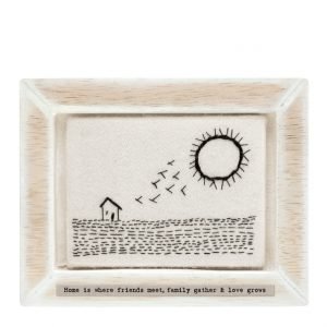 A wooden frame which is fitted around a piece of felt which is embroidered with an image of a house, field, birds and the sun on it. Printed on the frame underneath the felt are the words 'Friends meet, family gather and love grows'