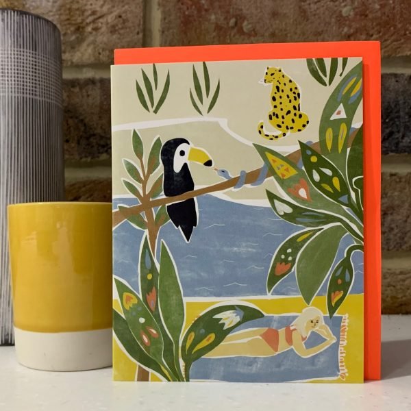 A blank greetings card with an image of a woman lying on a twoel next to a river which is in the jungle with toucan and a snake on a branch and a leaopard on the other side of the river.