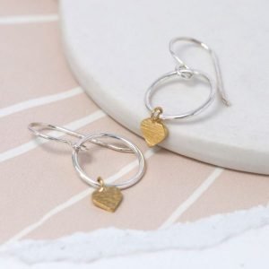 A pair of sterling silver drop earrings which have a hoop design with a scratched gold plated heart on the bottom of it.