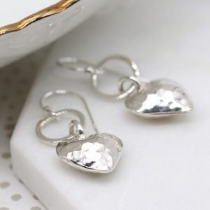 A pair of silver drop earrings with a hammered heart hanging from the loop.
