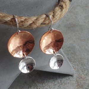 A pair of hand crafted earrings made from copper and sterling silver. Two hammered texture discs suspended from a silver ear wire. The top disc is larger (2cm) and is made of copper, the bottom disc is smaller (1.2cm) and made of silver.