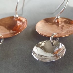 A pair of hand crafted earrings made from copper and sterling silver. Two hammered texture discs suspended from a silver ear wire. The top disc is larger (2cm) and is made of copper, the bottom disc is smaller (1.2cm) and made of silver.