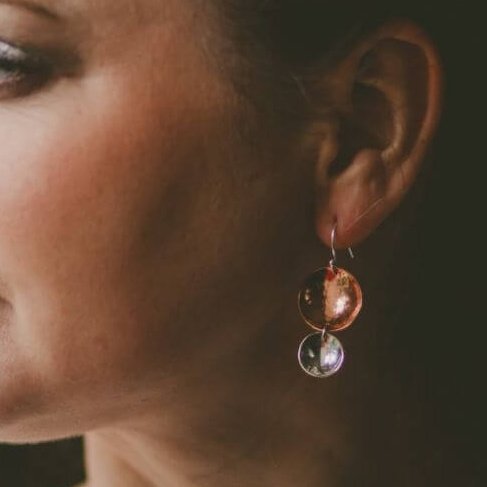 A pair of hand crafted earrings made from copper and sterling silver. Two hammered texture discs suspended from a silver ear wire. The top disc is larger (2cm) and is made of copper, the bottom disc is smaller (1.2cm) and made of silver.