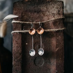 A pair of hand crafted drop earrings in sterling silver and copper. A silver ear wire with a hammered copper disc from with a silver wire extends to hold another hammered silver disc.