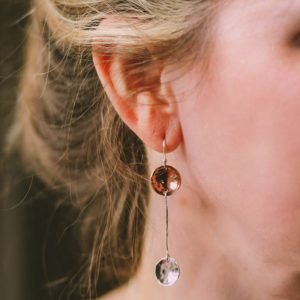 A pair of hand crafted drop earrings in sterling silver and copper. A silver ear wire with a hammered copper disc from with a silver wire extends to hold another hammered silver disc.