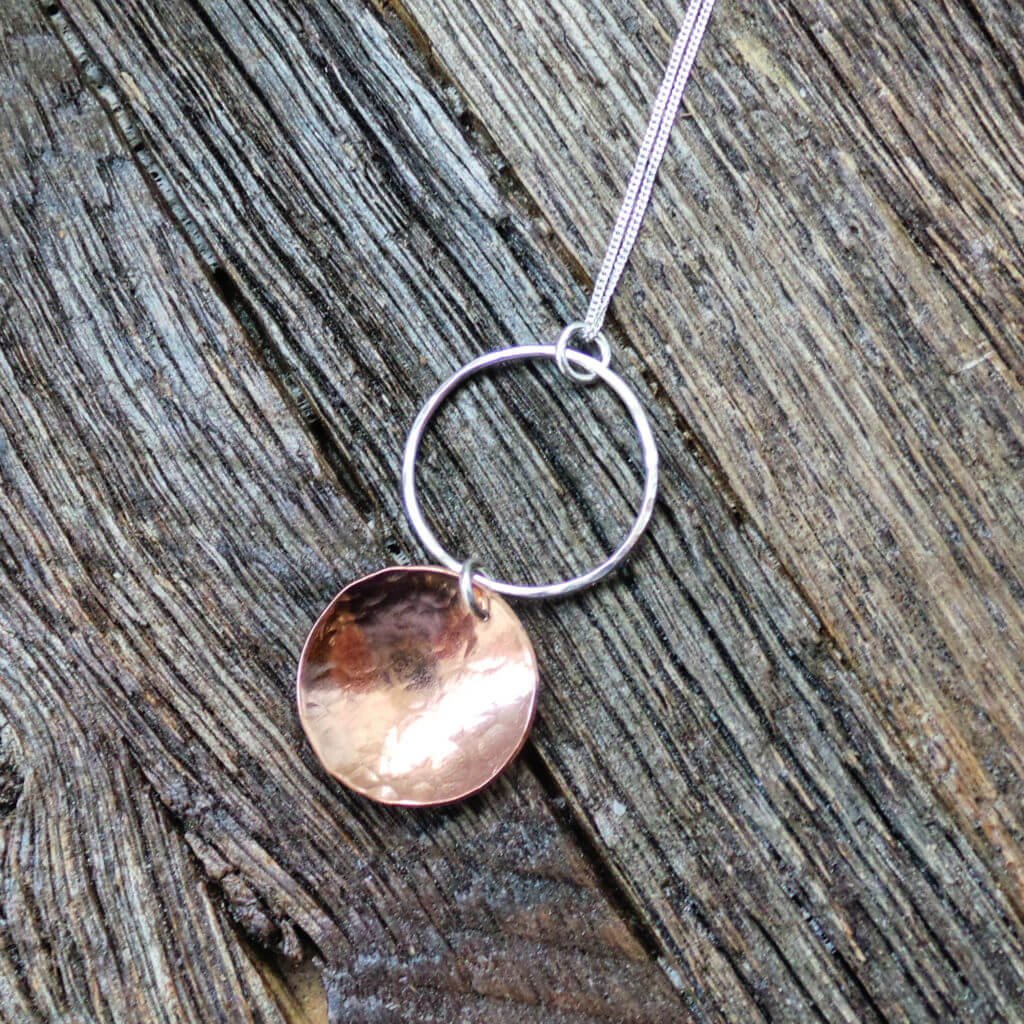 A hand crafted silver and copper mixed metal necklace. A silver hammered circle of silver with a hammered textured copper disc suspended from it. Comes in a gift box