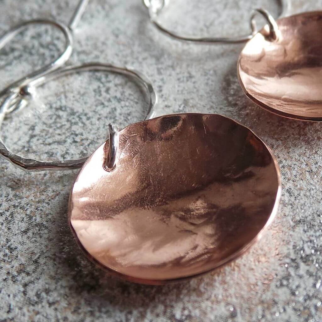 A pair of hand crafted silver and copper mixed metal earrings. A silver ear wire holds a hammered circle of silver with a hammered textured copper disc suspended from it. The earrings have approximately 5cm drop and come in a gift box