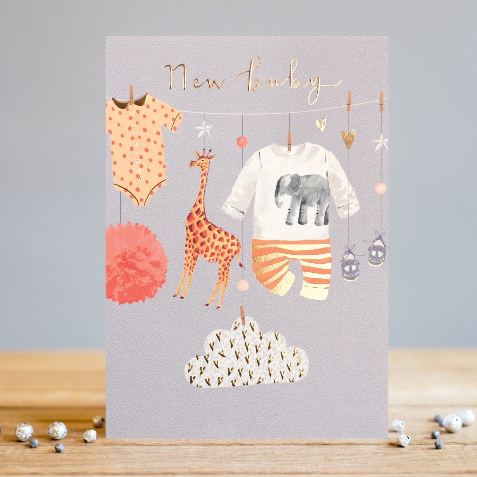 A sweet card from Louise Tiler. A neutral New Baby Card with a pale grey background. The image on the card is of new baby clothes hanging on a washing line along with a cute giraffe toy. The words New Baby are embossed, foiled and printed at the top of the card.