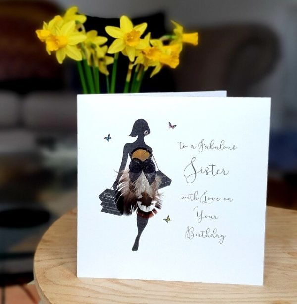 A handmade birthday card for a sister's birthday. A silhouette of a woman doing a sassy walk with a handful of shopping bags and a dress made of real feathers. The card has little butterflies and crystals and the words To a fabulous Sister with love on your birthday.