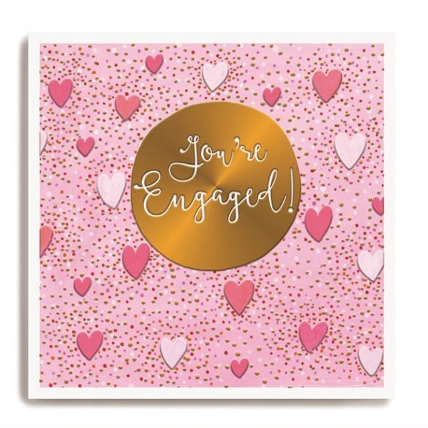 A square card with a pink background which is covered in dots and hearts. In the centre of the card is a gold disc which has been embossed with the words You're Engaged on it.