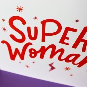 A square white card with the words 'Super Womam' printed in vibrant red, with images of stars and lightening bolts around it.