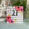 A square card with a grey striped background with colourful flowers in the corners and little gold spots on it. The number 21 is printed in the centre of the card in black.