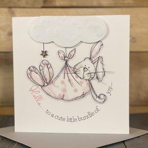 A lovely Hello little bundle of joy card with an image of a rabbit being carried in a pink blanket below a fluffy cloud.