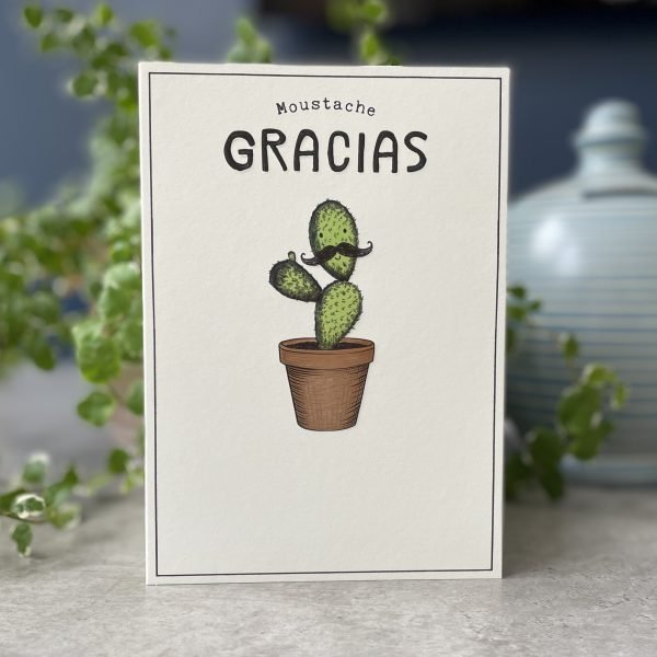 A cream coloured card with an image of a cactus in a pot with a face on the top section . The face has a curly moustache on it. The words Moustache Gracias are printed above the cactus image.