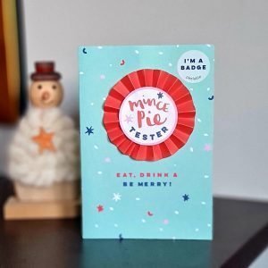 A christmas card with a paper rosette style removable badge that says Mince Pie Tester