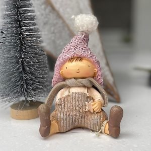 A cute sitting boy made from ceramic with beige coloured trousers. A beige scarf, Pink knitted style hat with white pompom on top.