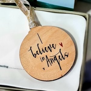 A wooden disc painted with an Angel of the North in silhouette on a gold landscape. the reverse of the disc says I believe in angels