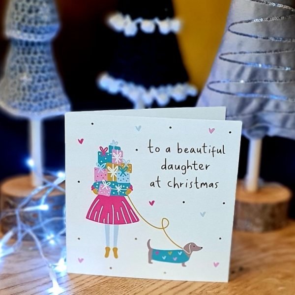 A Christmas card with a brightly coloured illustration of a girl holding a pile of gifts holding a sausage dog on a lead. To a beautiful daughter at Christmas in gold foil text
