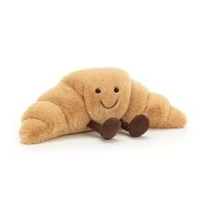 A cute cuddly toy made to look like a croissant with a cute smiley face and little chocolate coloured legs and feet.