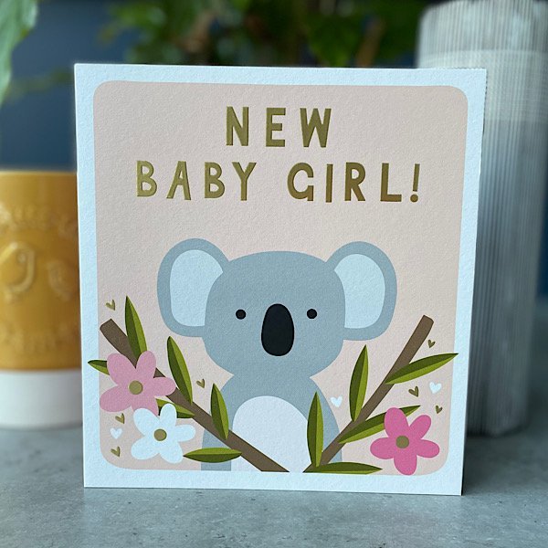 A square card with an image of a Koala with plants around with and the words New Baby Girl embossed and printed on it,