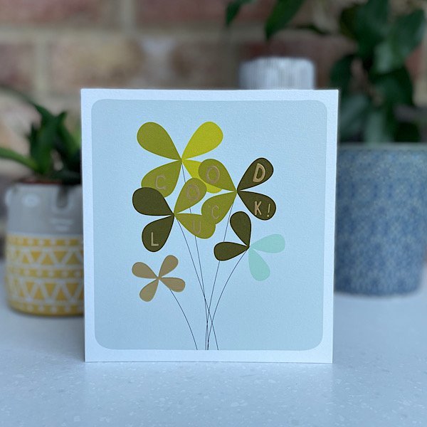 A square card with an image of a bunch of four leaf clovers and the words Good Luck printed on the leaves.