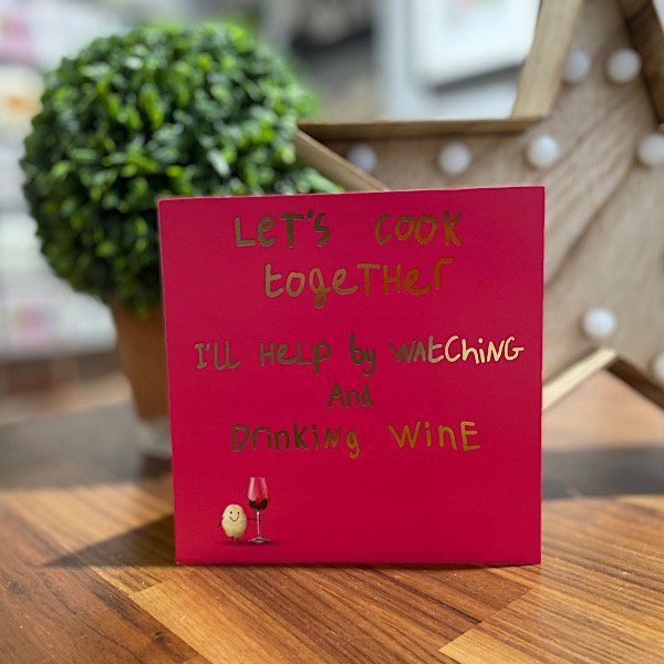 A burgundy card with Let's cook together. I'll help by watching and drinking wine printed in gold foil