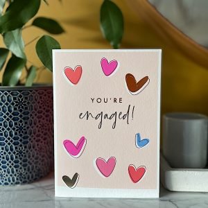 A pink coloured card with colourful hearts all over it and the words you are engaged printed on it.