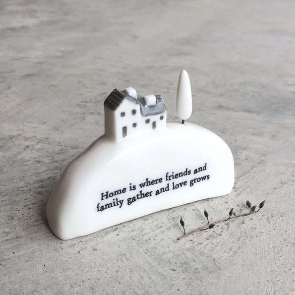 A lovely ceramic ornament with a sweet house and tree on it. The words Home is where friends and family gather and love grows. on the hill area of the ornament.