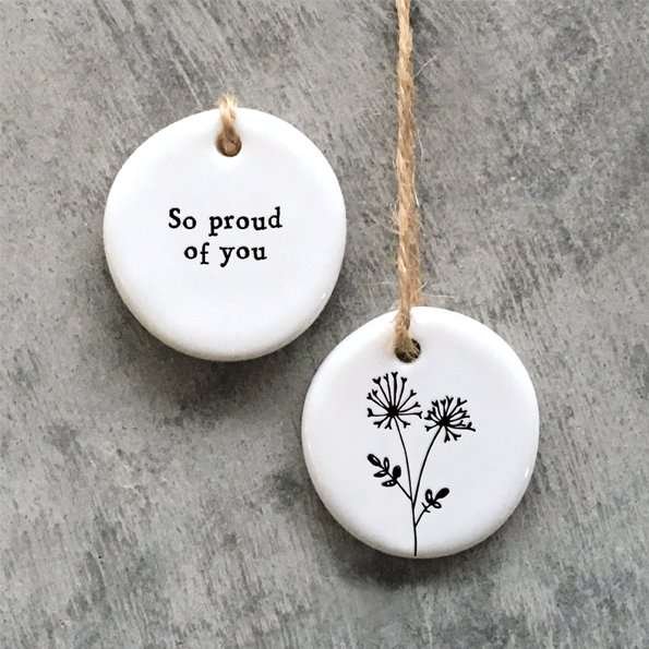 A ceramic pebble keepsake with a little floral design on one side and the words So proud of You on the other side. The pebble has a brown string hanger.
