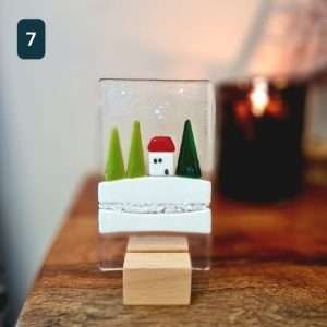 A fused glass Christmas ornament with trees, snow and a little house. On a wooden stand