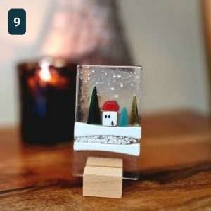 A fused glass Christmas ornament with trees, snow and a little house. On a wooden stand