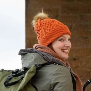 An burnt orange fleece lined, cable knit bobble hat in wool mix with a natural fuax fur pom pom